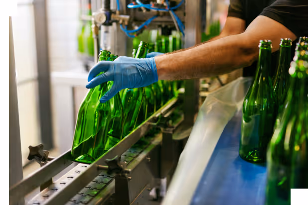Worker wearing a blue glove handling green glass bottles on a conveyor belt in a bottling plant.