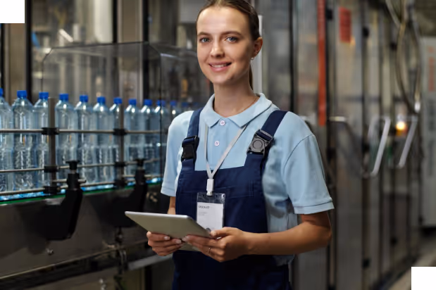 Woman in work uniform holding a tablet standing next to a conveyor belt with bottled water in a factory.