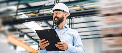 Smiling construction manager wearing a white hard hat holding a clipboard inside an industrial building.
