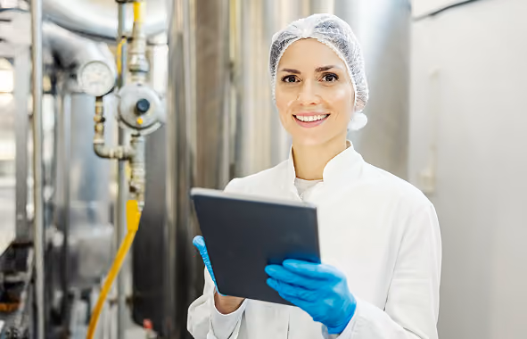 Smiling plant manager in white coat and hairnet holding tablet in industrial facility.