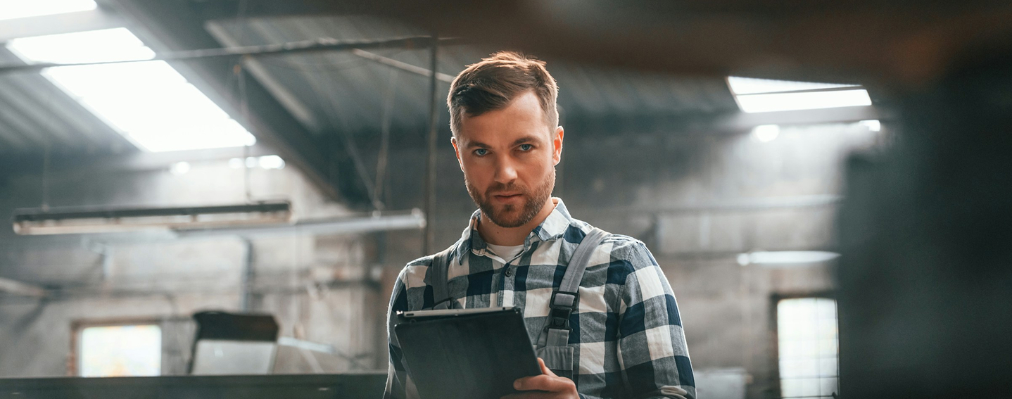 Man in checkered shirt holding a clipboard in an industrial warehouse setting.