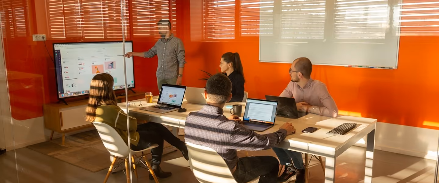 Five people in a meeting room; one is presenting data on a large screen, others sit with laptops.