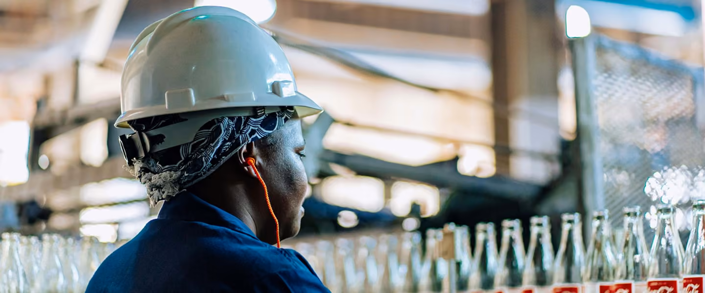 Worker with a white hard hat and earphones in a factory near rows of glass bottles.