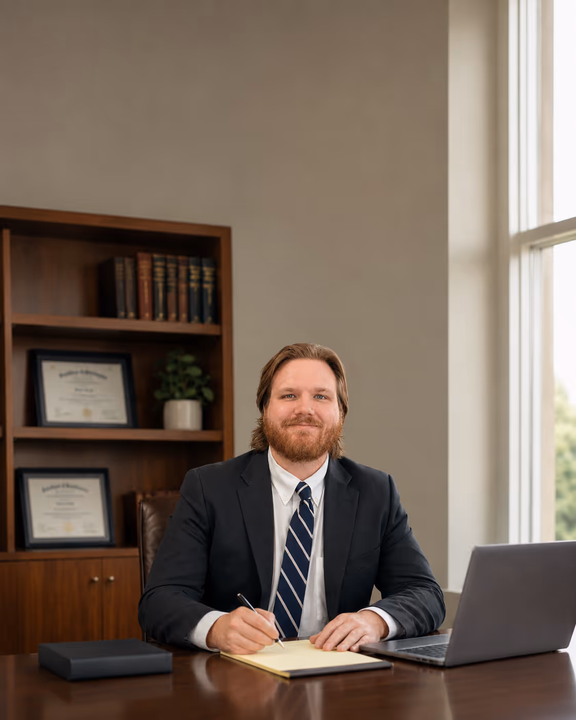 Smiling man in a suit and tie writing on a yellow notepad at a desk with a laptop and bookshelf behind.