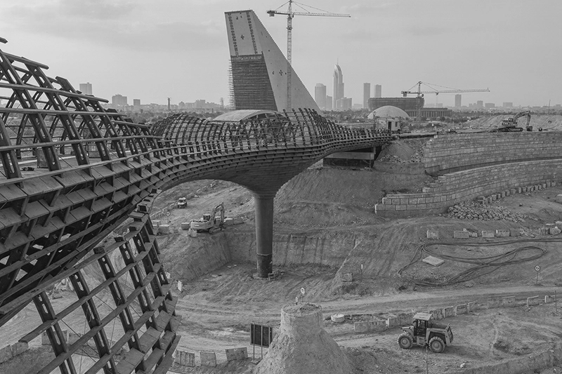 Wooden lattice bridge under construction in a large excavation site with cranes and buildings in the background. King Salman Park, Riyadh, KSA.