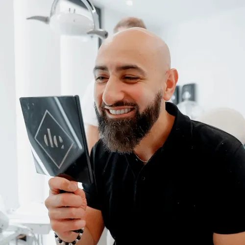 A man with a beard, smiling into a  hand held mirror.