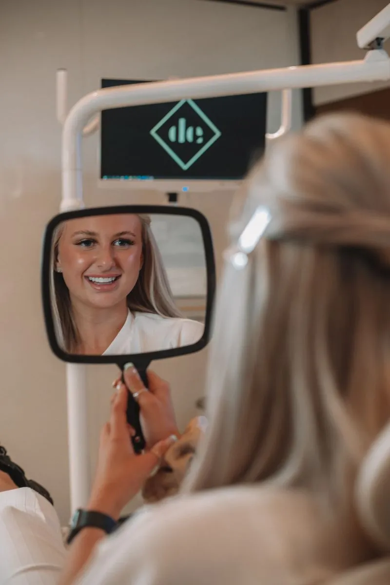 A woman in a white shirt smiling into a handheld mirror.