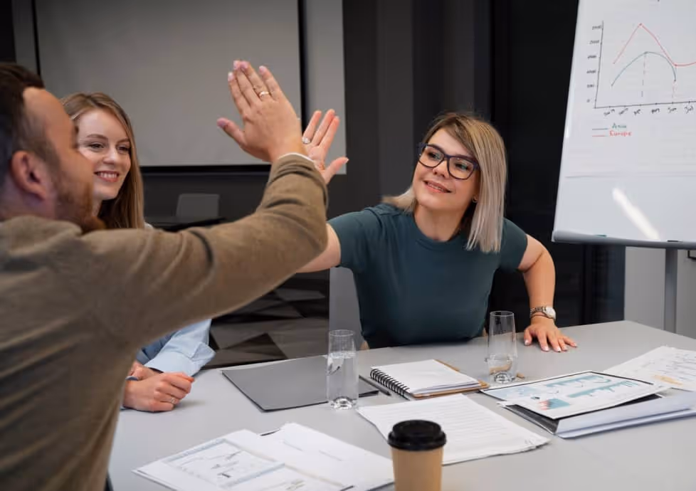 Three coworkers at a meeting table giving a high five with papers and a whiteboard chart nearby.