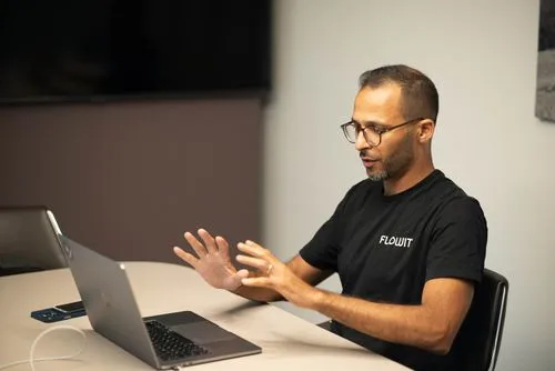 Man wearing glasses and Flowit shirt sits at desk gesturing toward laptop screen.