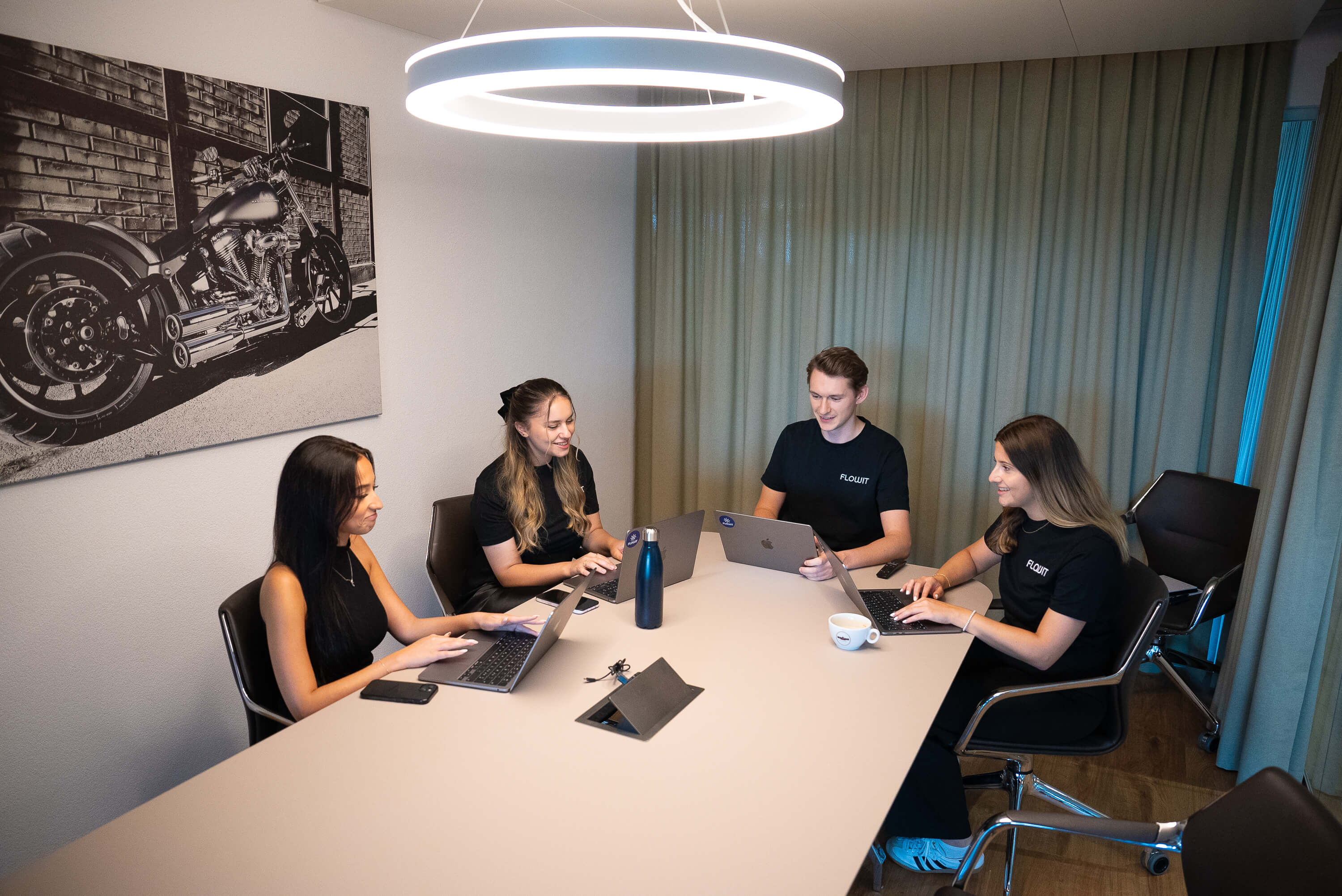 Four people seated around a table, working on laptops in a modern meeting room with a motorcycle photo on the wall.
