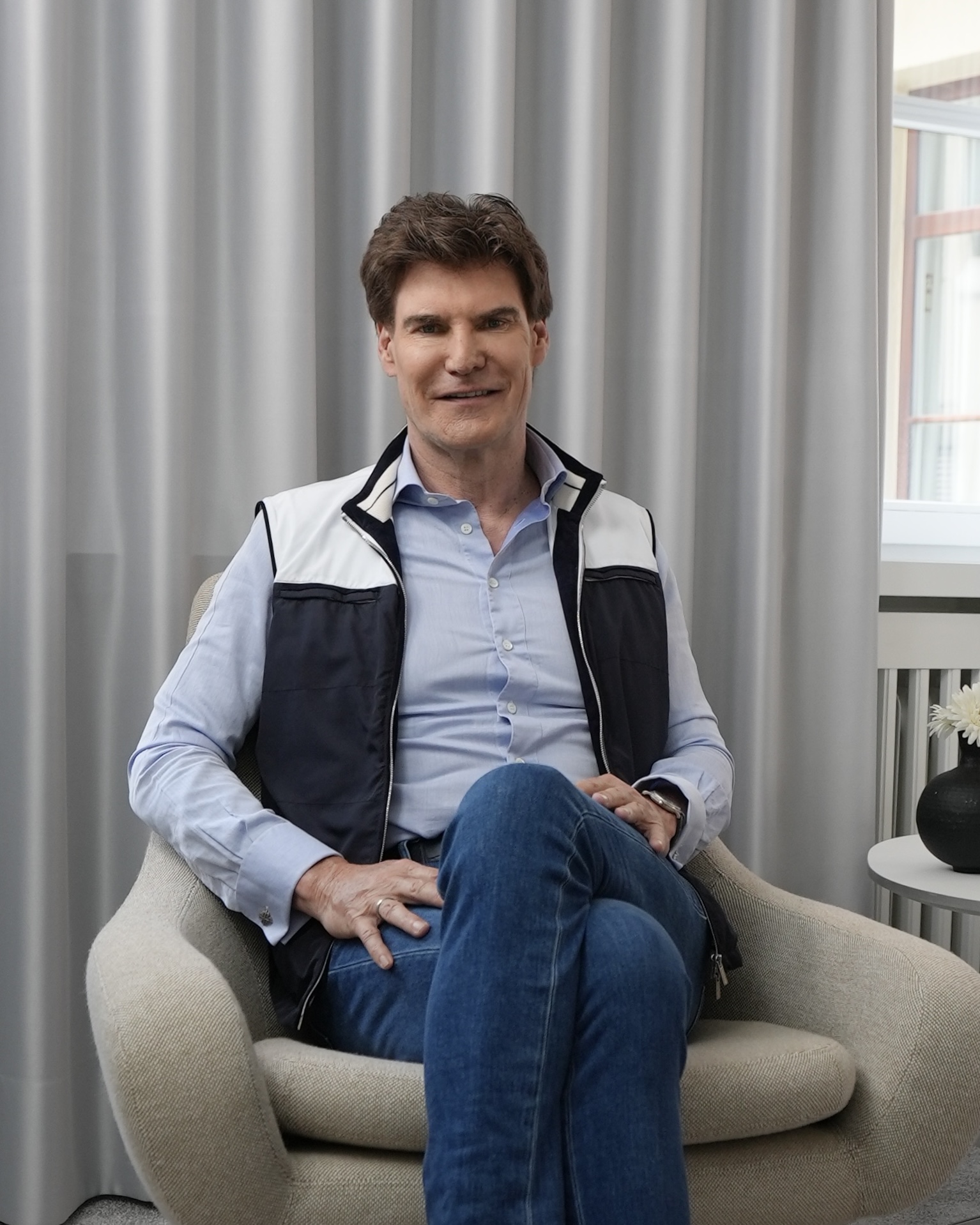 Man with brown hair wearing a blue shirt and vest sitting cross-legged in a beige armchair.