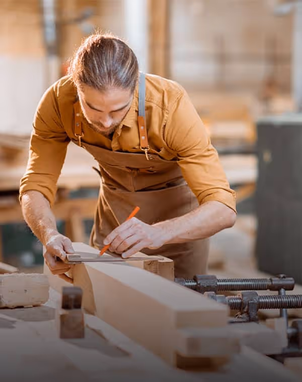 Carpenter wearing apron marking a wooden plank with a pencil in a woodworking shop.
