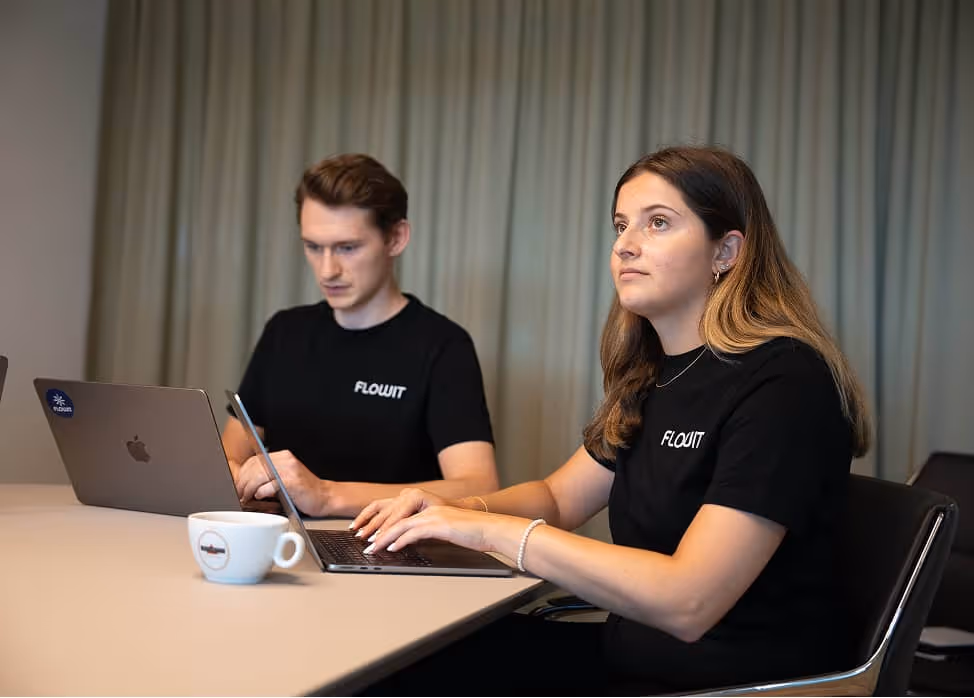 Two people wearing black FlowIt shirts working on laptops at a table with a coffee cup in front.