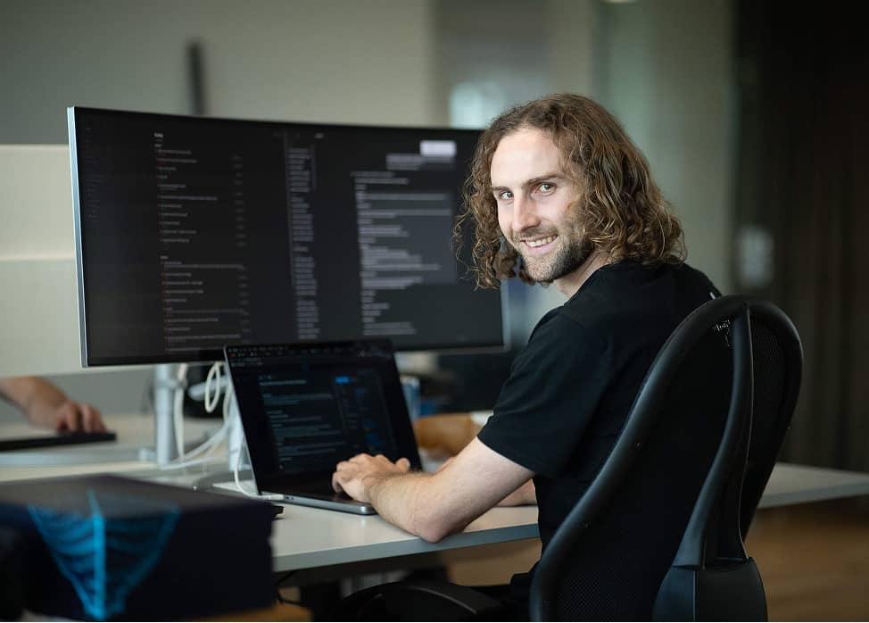 Man with curly hair smiling while working on laptop with large monitor displaying code in office.