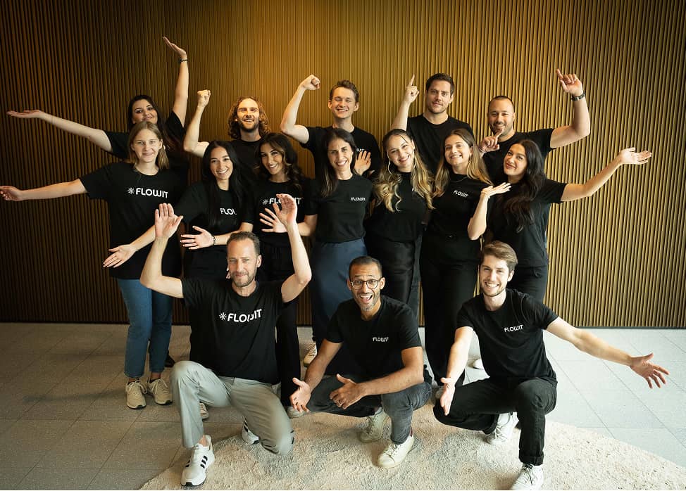 Group of 15 people in black FLOWIT shirts posing happily with raised arms indoors.