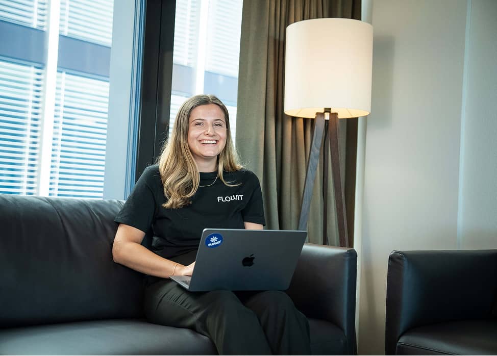 Smiling woman in black FLOWIT shirt sitting on sofa with a laptop in an office setting with lamp and window.