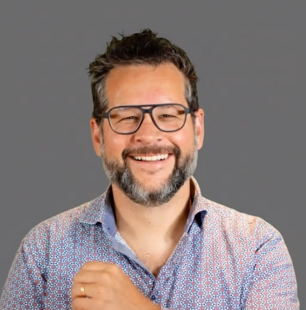 Smiling man with glasses, beard, and patterned shirt in front of a gray background.
