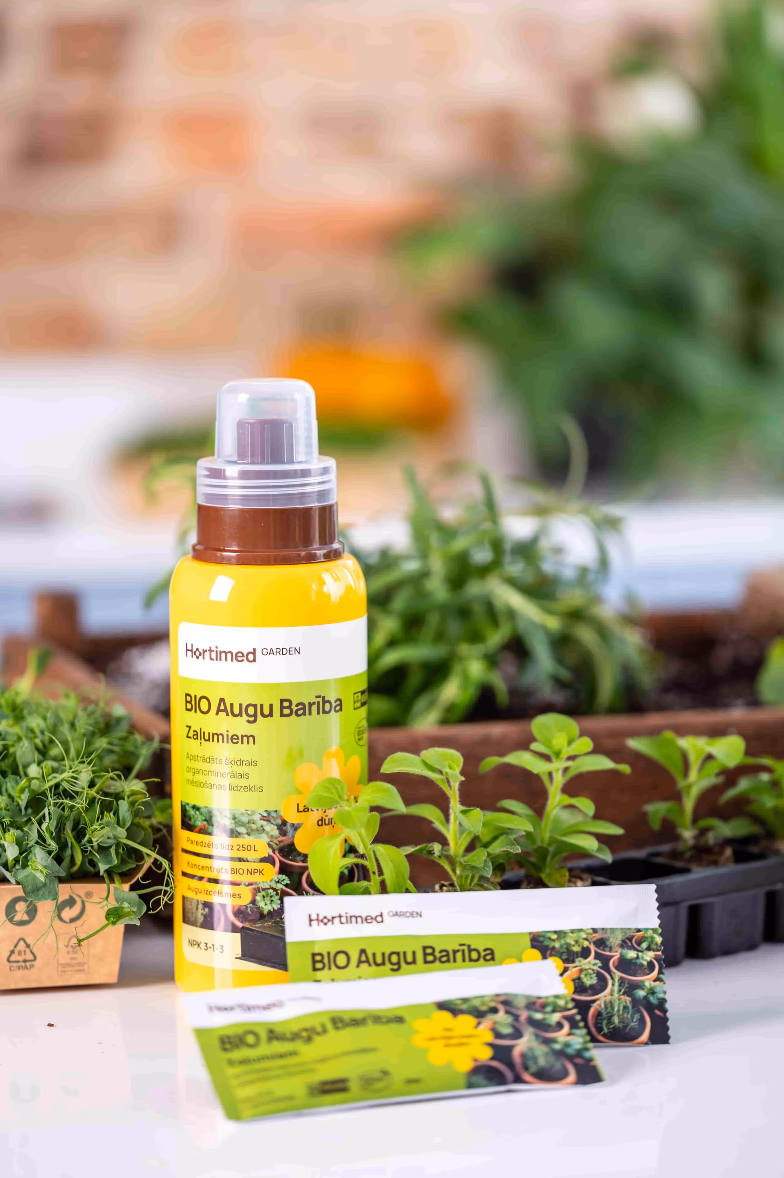 Yellow bottle of Hortimed Garden BIO Augu Barība plant fertilizer surrounded by green seedlings and small potted herbs.