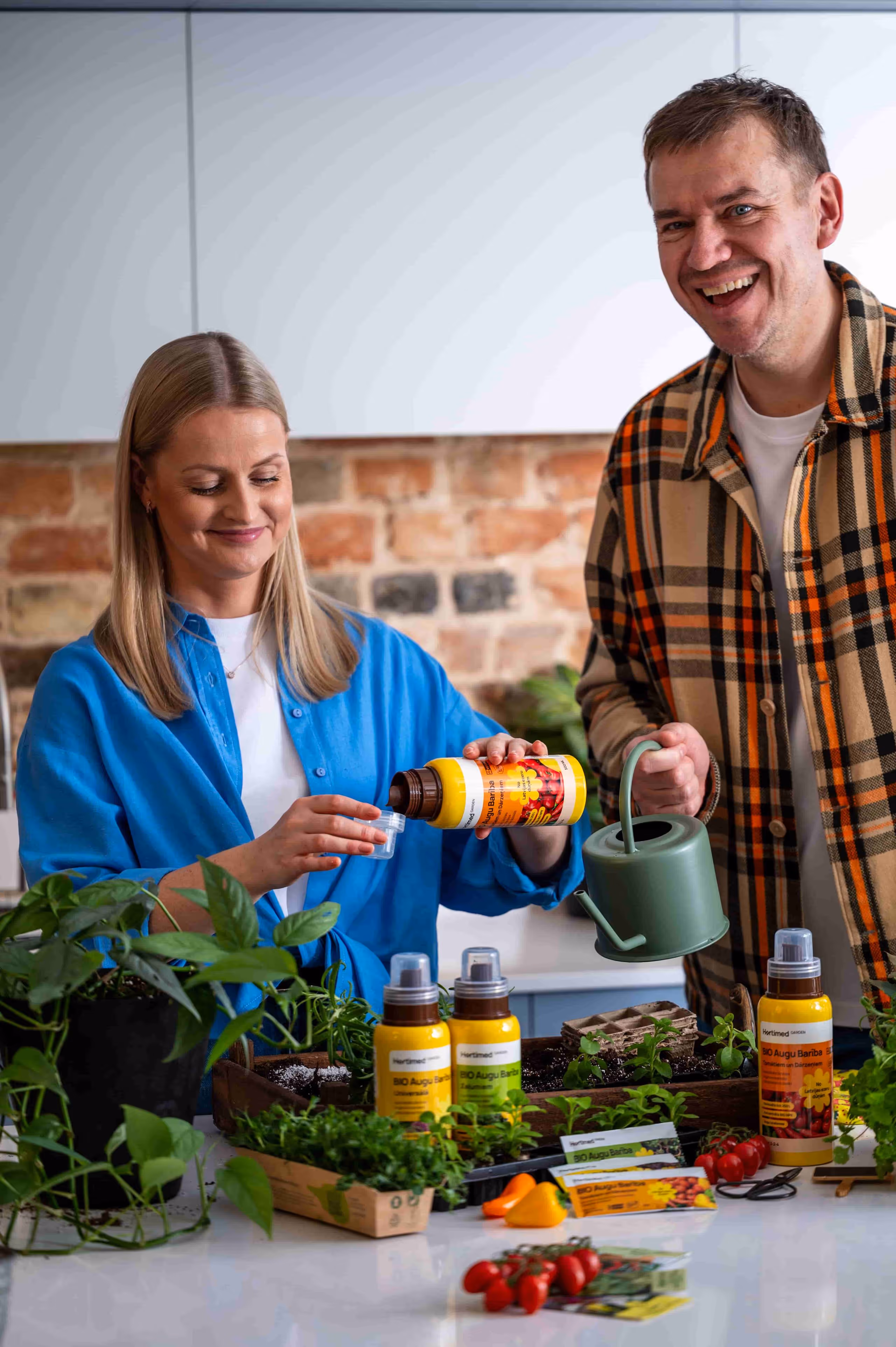 Woman pouring liquid plant fertilizer into a small cap while a man holds a green watering can, surrounded by various plants and gardening supplies on a kitchen counter.
