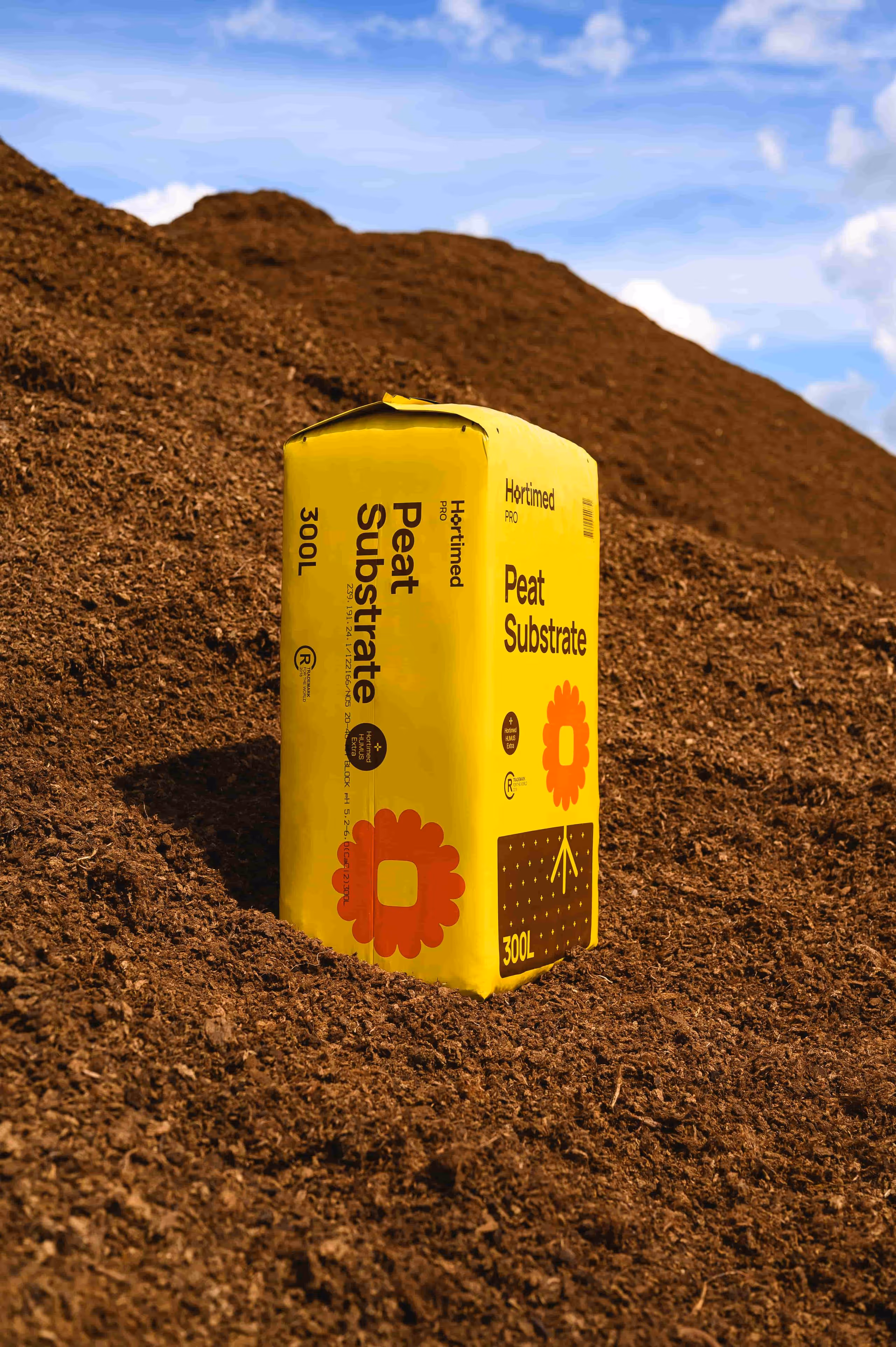 Yellow 300L bag of Hortimed Pro Peat Substrate standing on a large pile of brown soil under blue sky.