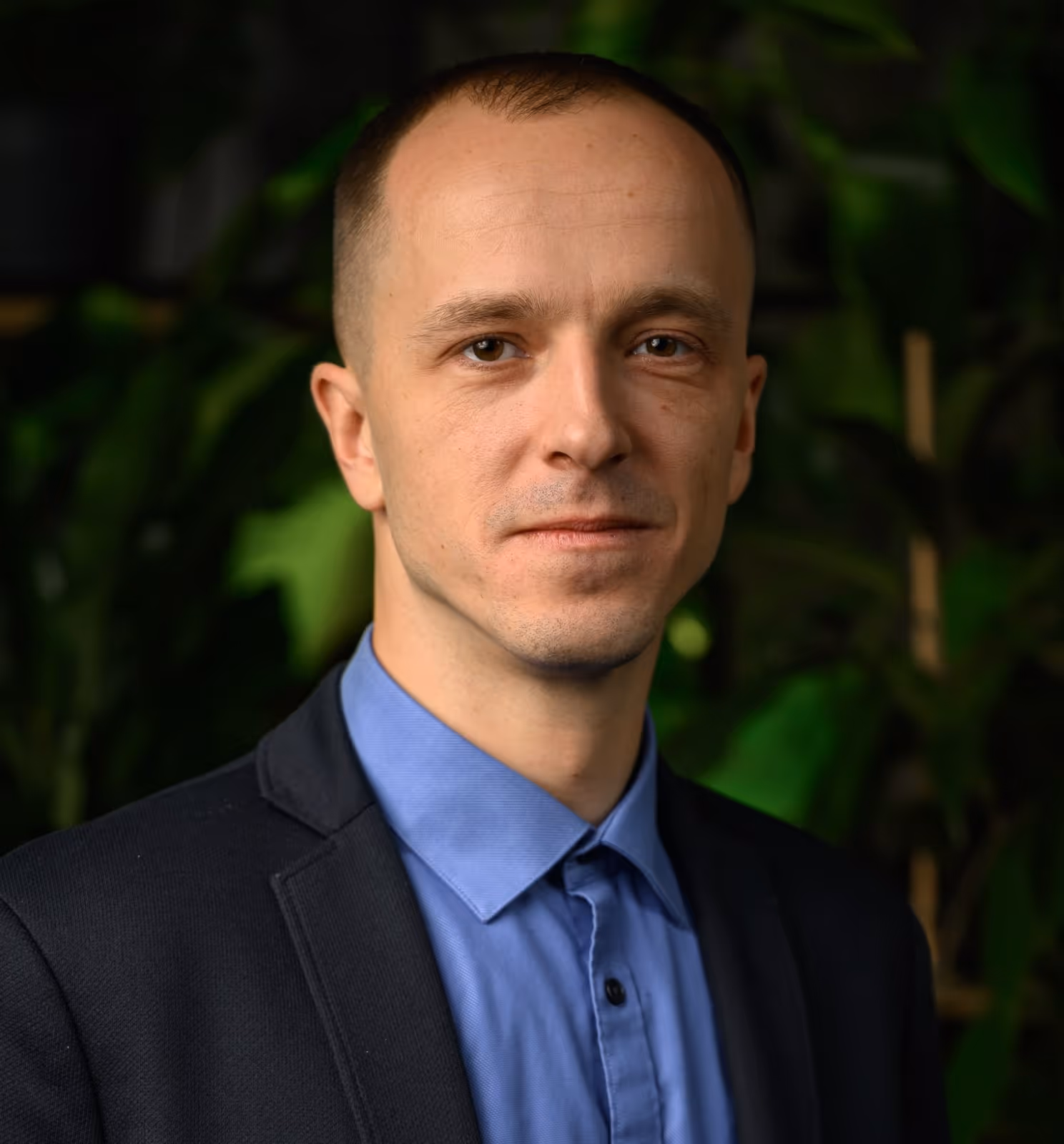 Portrait of a man in a blue shirt and black blazer with a blurred green leafy background.