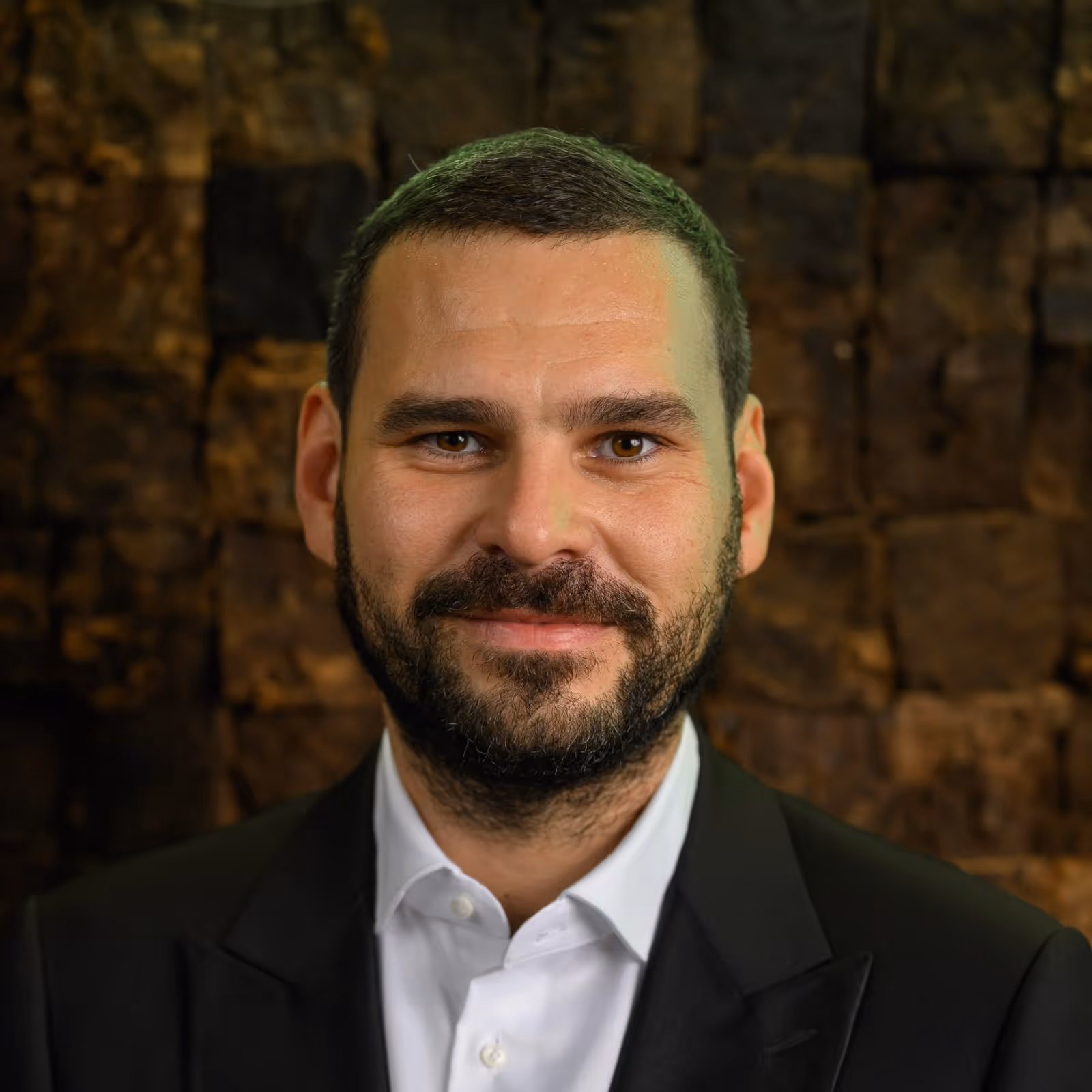 Smiling man with short dark hair and beard wearing a black suit and white shirt against a brown textured background.