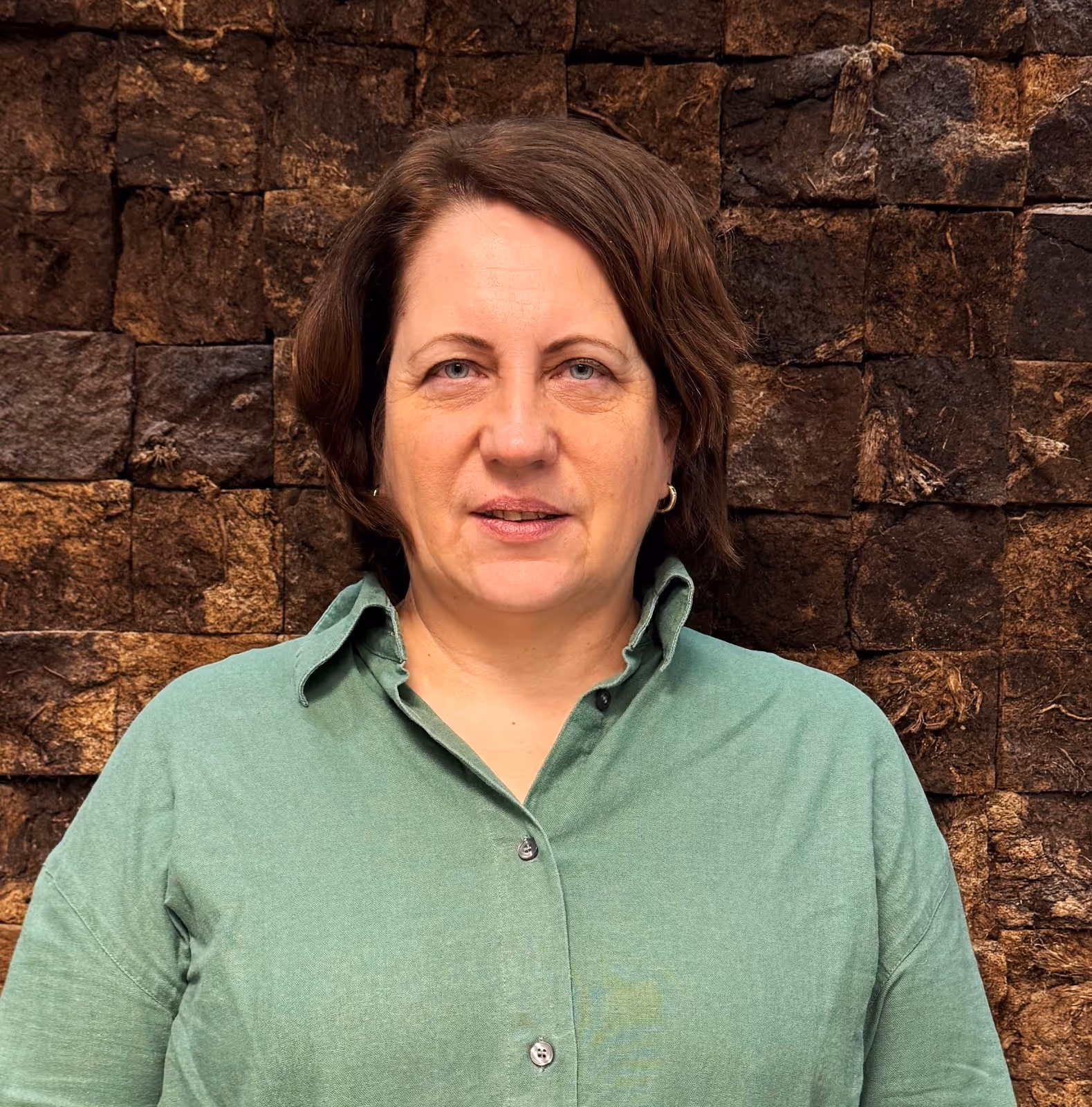 Middle-aged woman with short brown hair wearing a green collared shirt against a textured brown stone wall background.