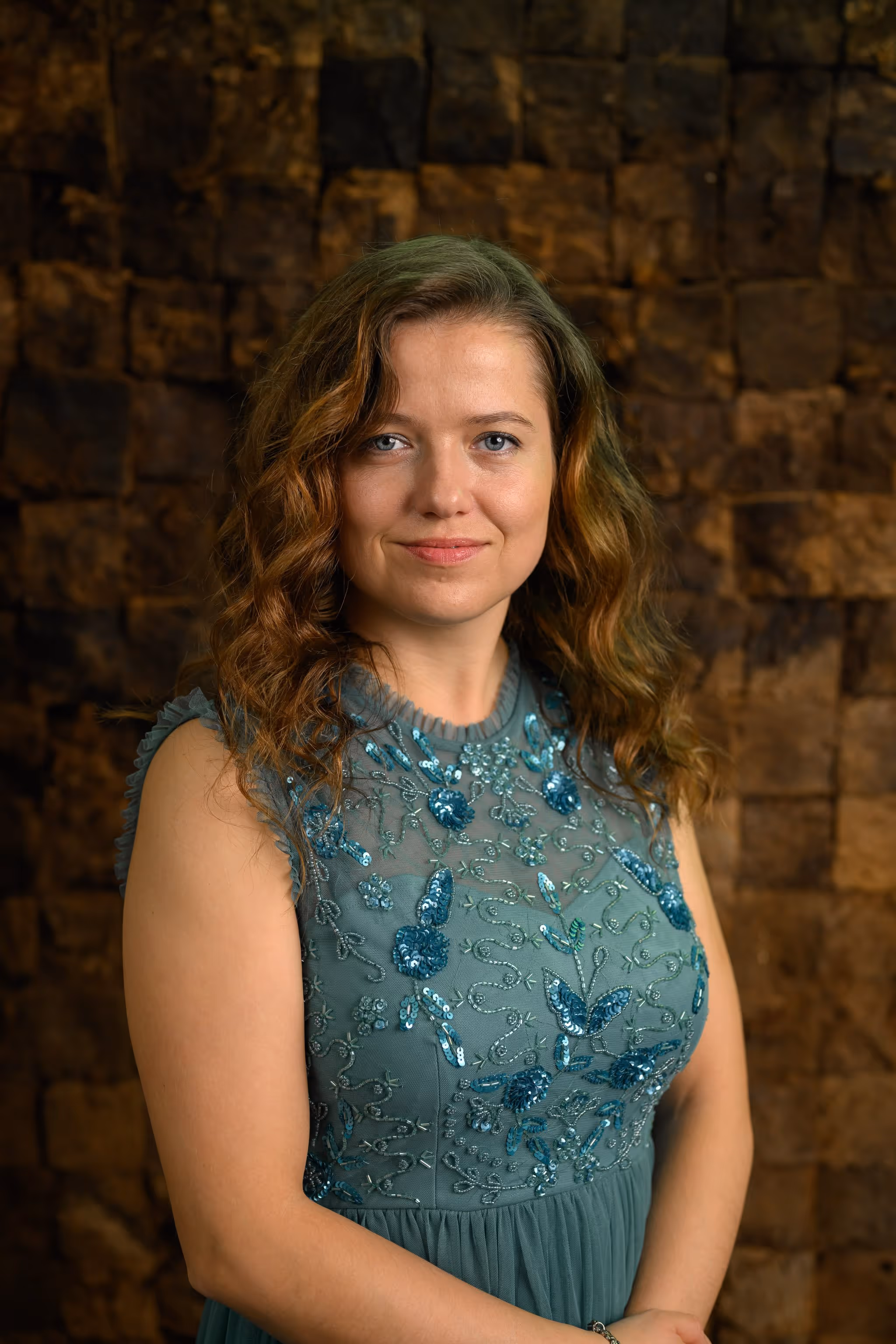 Woman with wavy light brown hair wearing a teal dress with sequined floral embroidery standing against a textured brown stone wall.