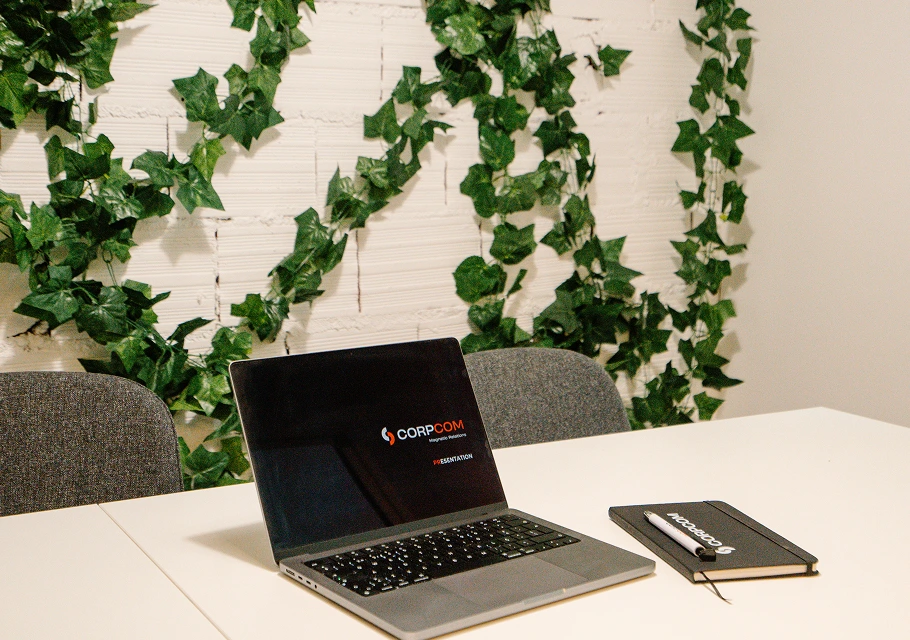 Computador portátil aberto sobre mesa branca com caderno e pen, fundo com parede branca e folhas verdes.