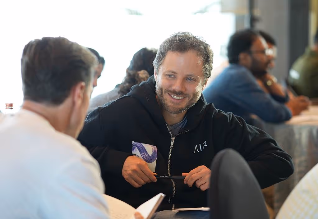 Smiling man in black hoodie holding a pen, engaged in conversation with another person at a table.