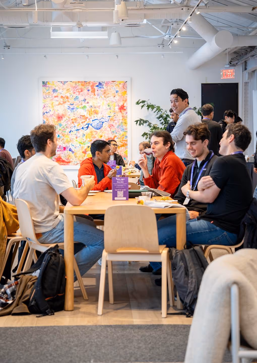 People sitting and standing in a bright modern room, talking and eating around tables with colorful artwork.
