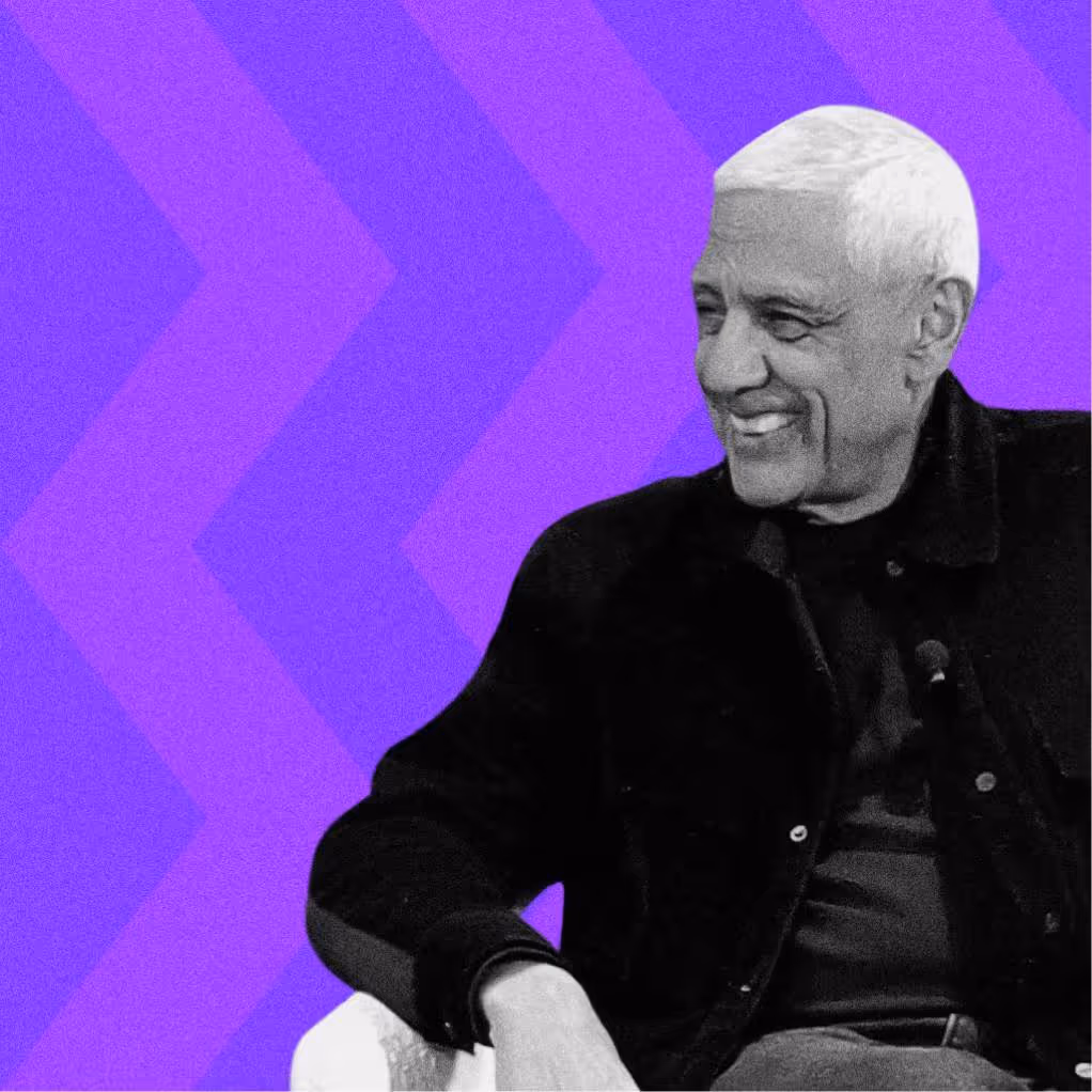 Smiling elderly man with white hair sitting against a purple geometric background.