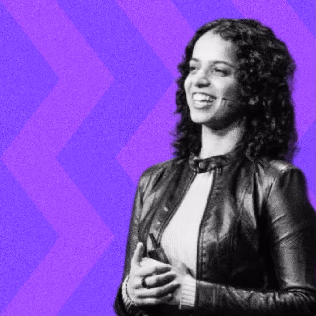 Smiling woman with curly hair speaking, wearing a leather jacket over a light sweater on purple background.