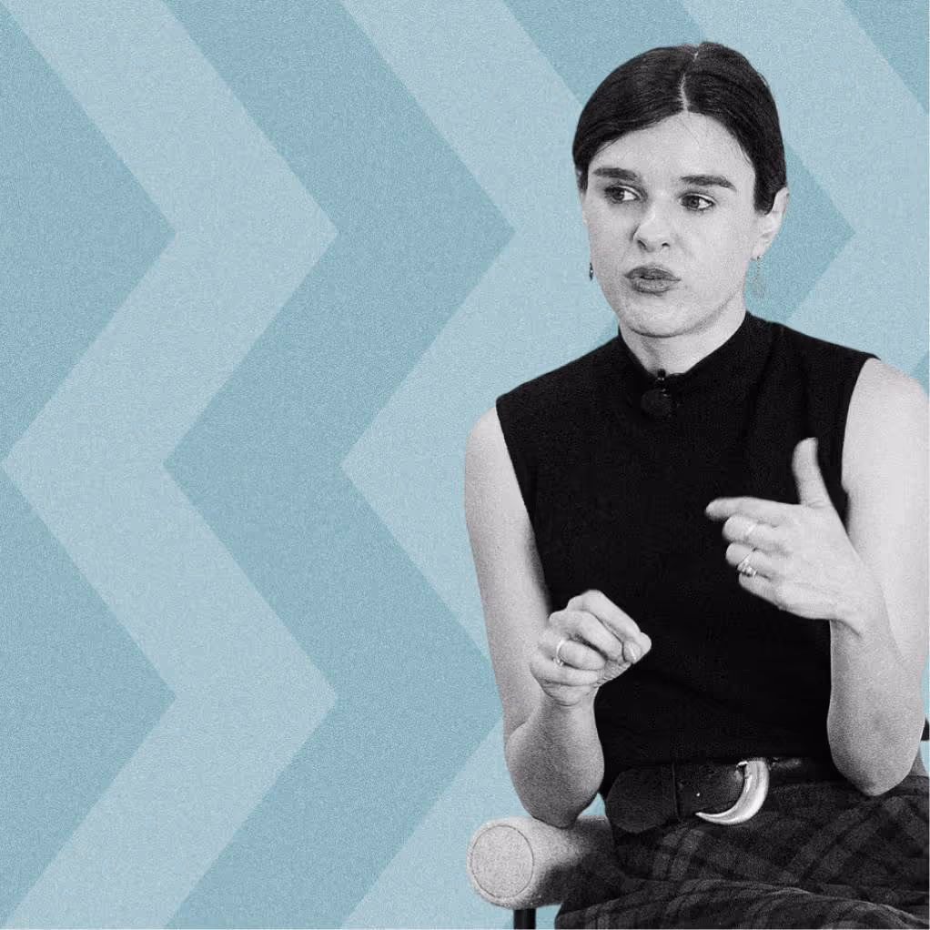 Woman in black sleeveless top with dark hair gestures while seated against a blue arrow-patterned background.