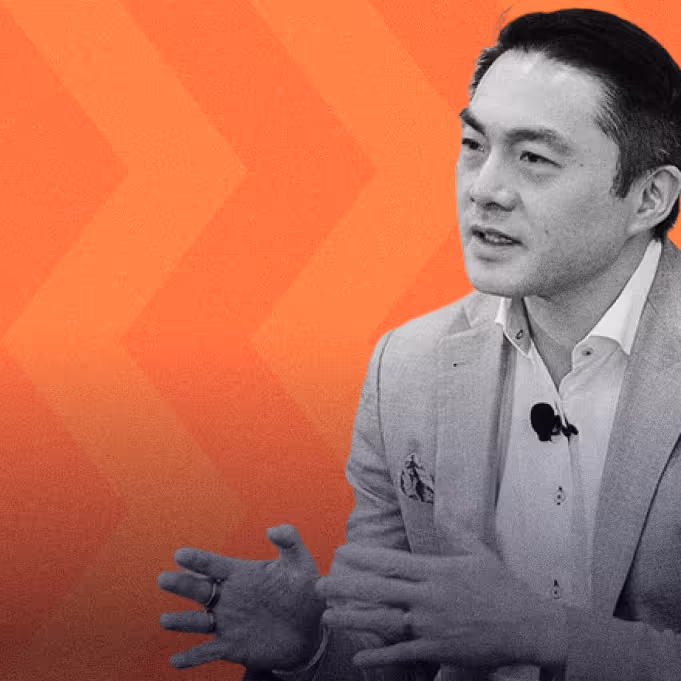 Man in a light blazer and white shirt speaking with hands gesturing on an orange chevron background.