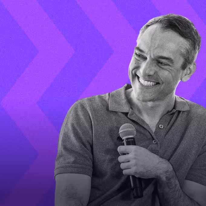 Smiling man in polo shirt holding a microphone against a purple background with chevron pattern.