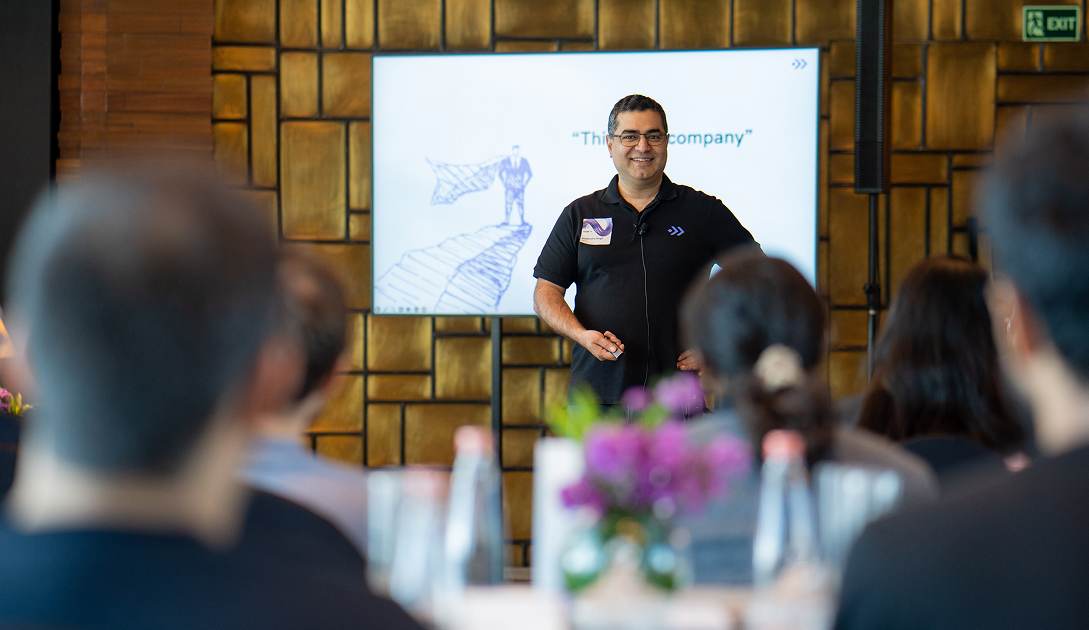 Man in black polo shirt giving a presentation to an audience with a screen behind him.
