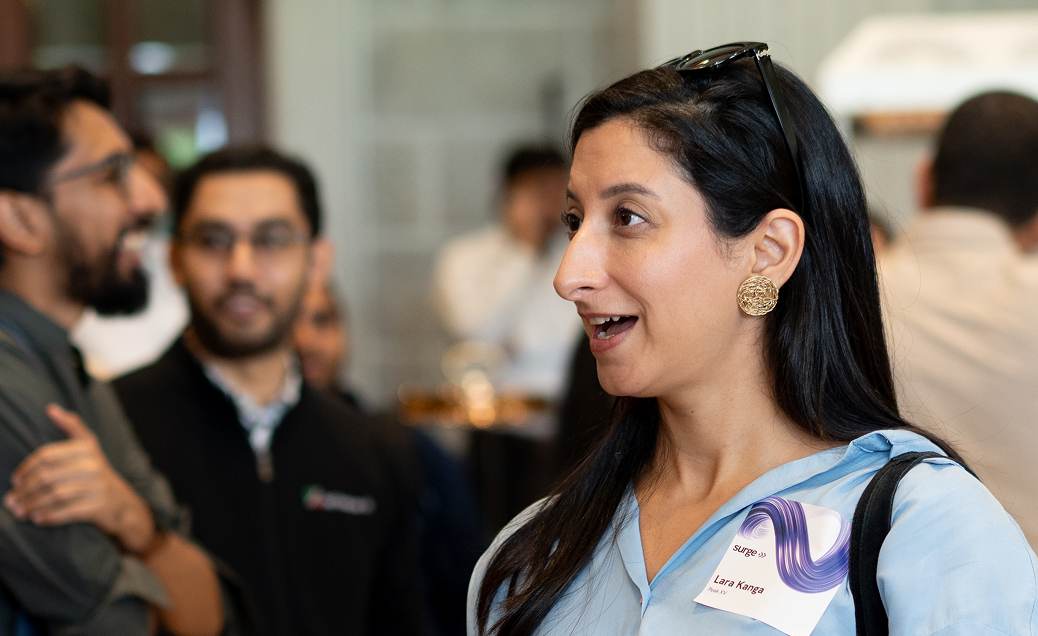 Woman with dark hair and gold earrings speaking at a social event with blurred background people.