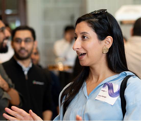 Woman in blue shirt with name tag talking to a group of people at an indoor event.