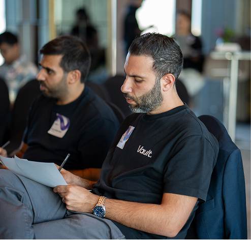Two men seated indoors, focused on reading papers, one wearing a black Vault t-shirt and watch.