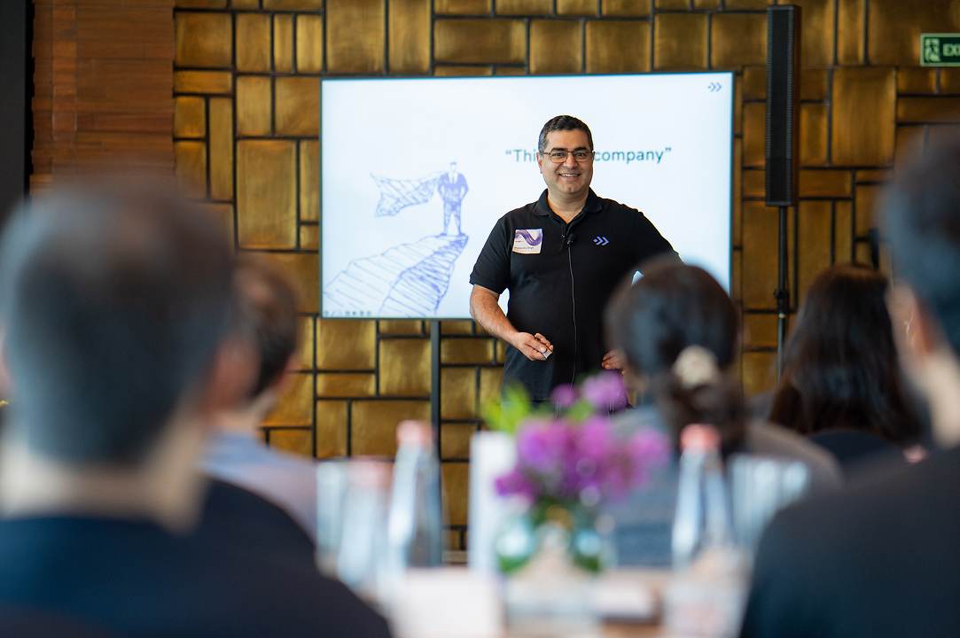 Smiling man giving a presentation to an audience with a slide behind him in a conference room.