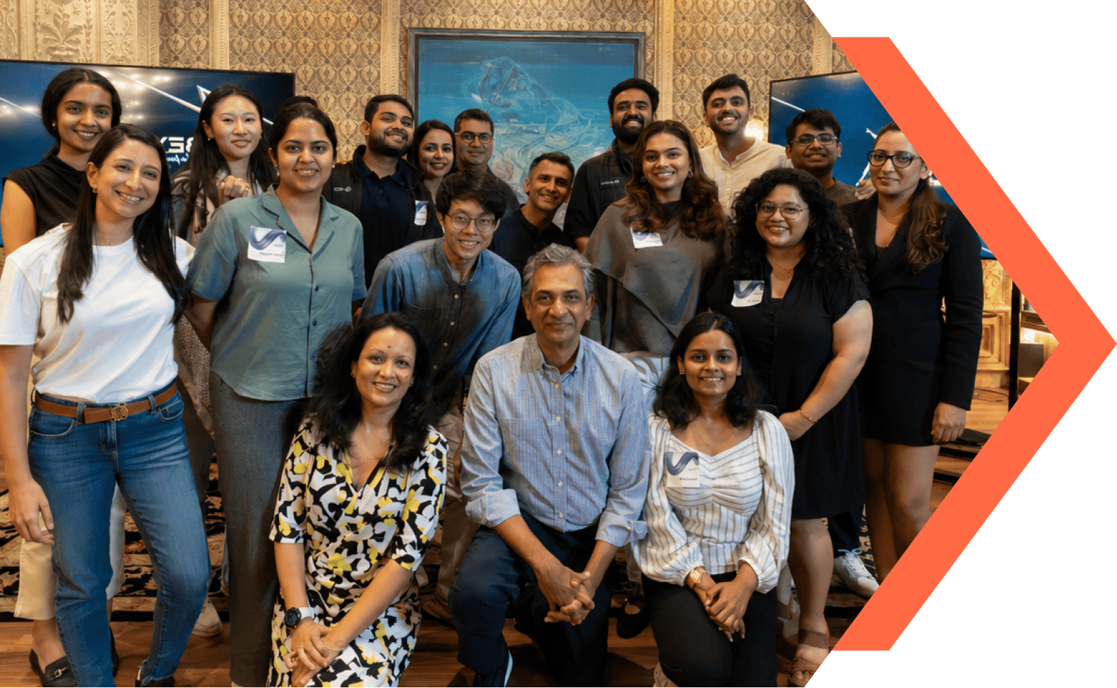 Group photo of a diverse team of 18 people smiling indoors in front of patterned wallpaper and screens.