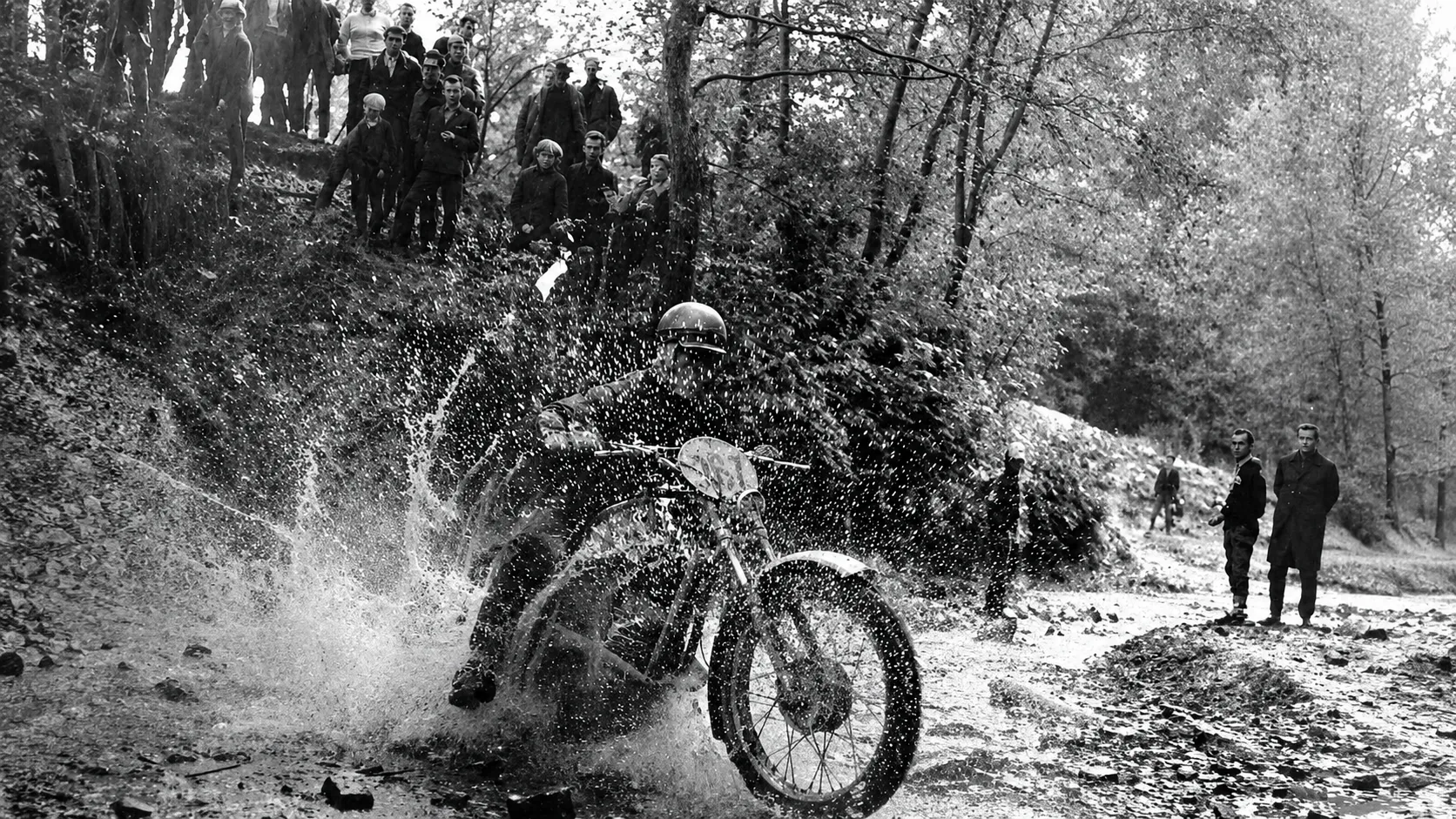 Black and white photo of a motorcyclist splashing through water while spectators watch in a wooded area.