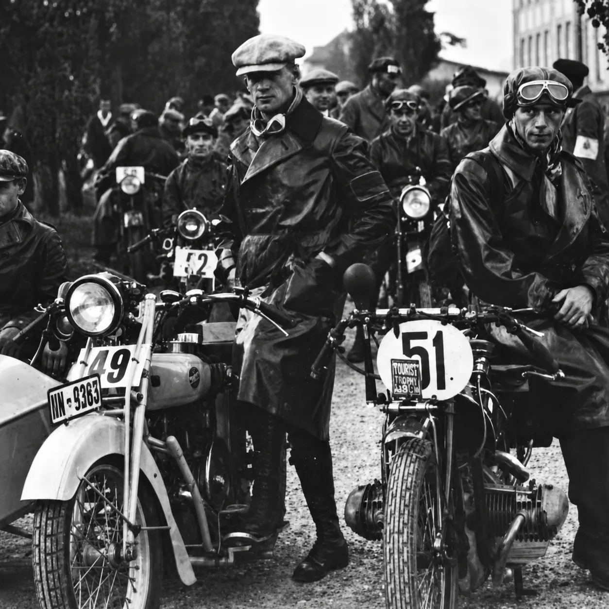 Black and white image of vintage motorcyclists in leather coats with numbered motorcycles, ready to race.