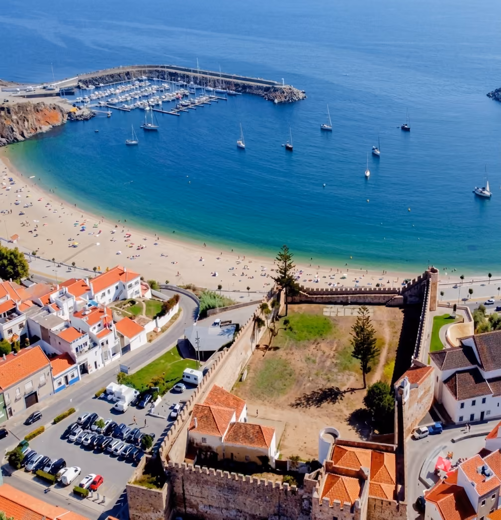 A coastal town with a beach, boats anchored in blue water, and a historic walled fortress.