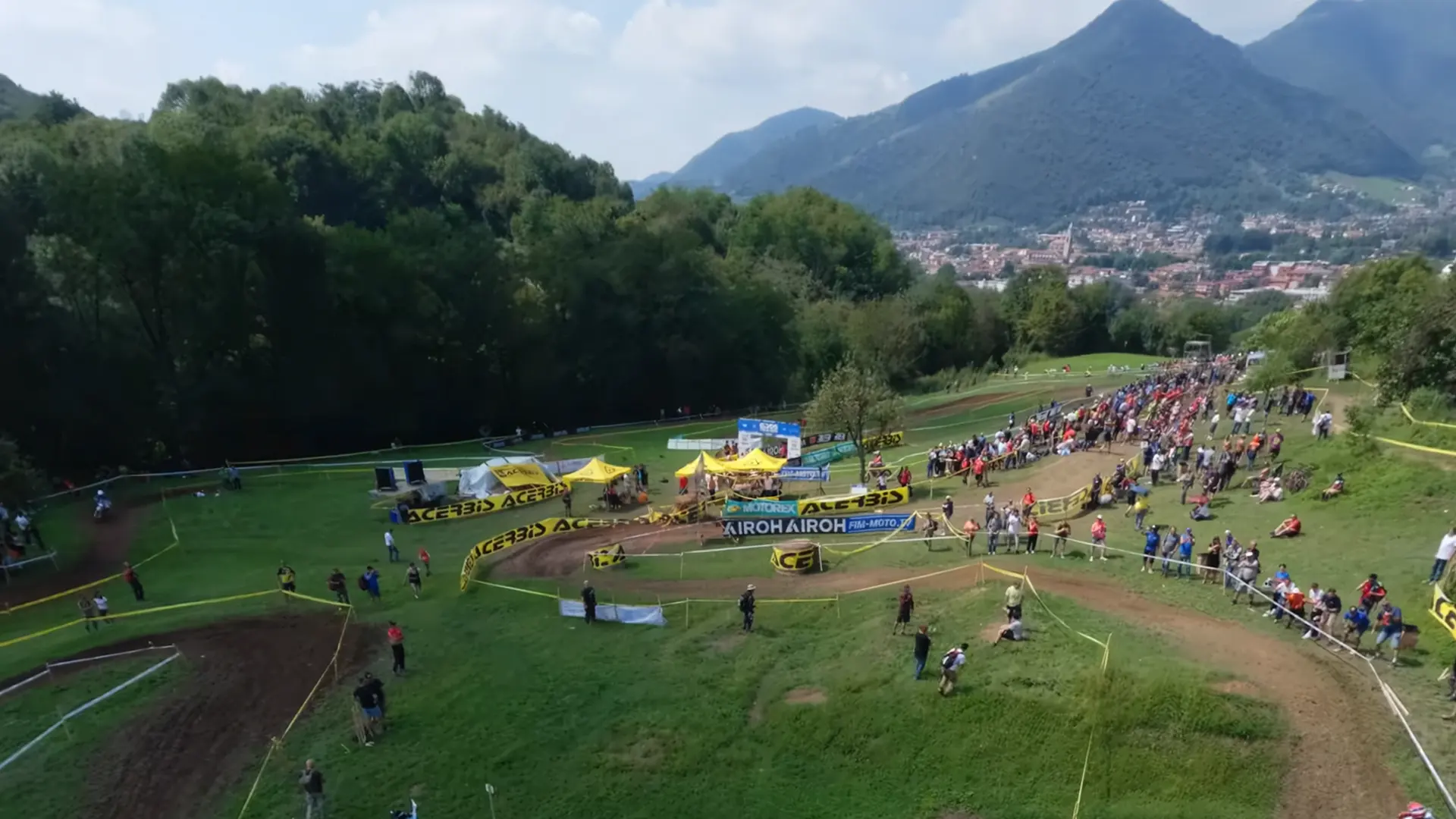 Outdoor motocross event with a dirt track, spectators, tents, and green hills in the background.