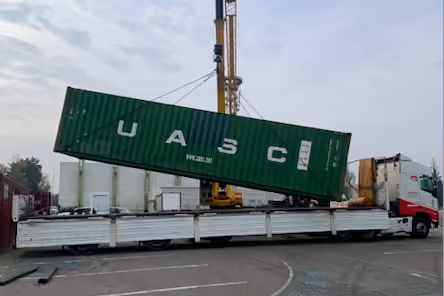 Green UASC shipping container being lifted onto a white flatbed truck by a crane.