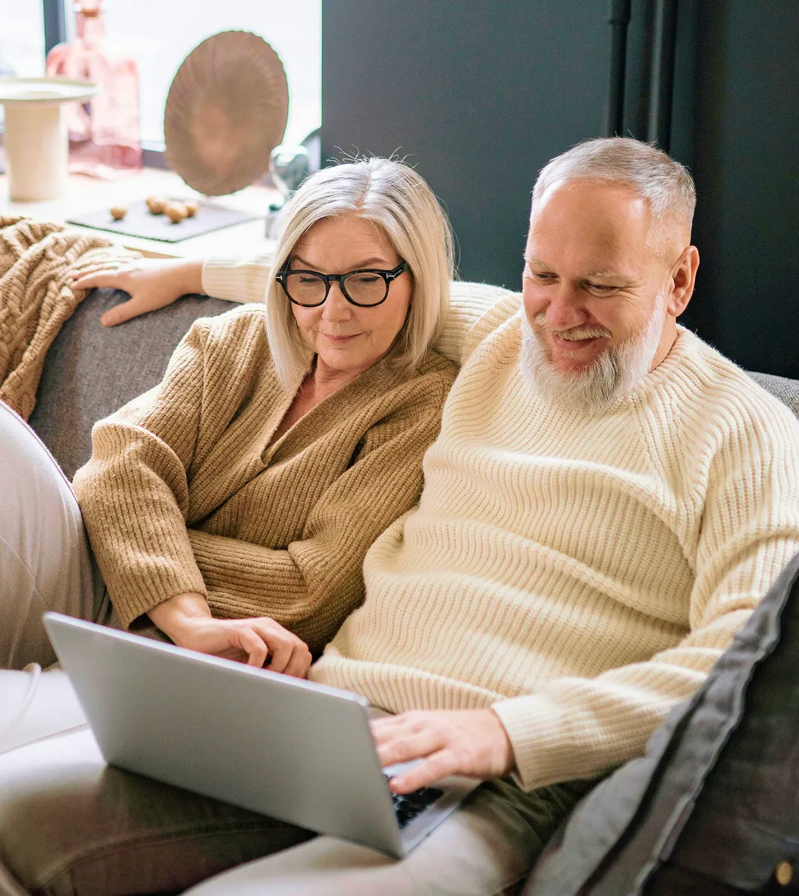 Older couple sitting on a couch together, looking at a laptop and smiling.