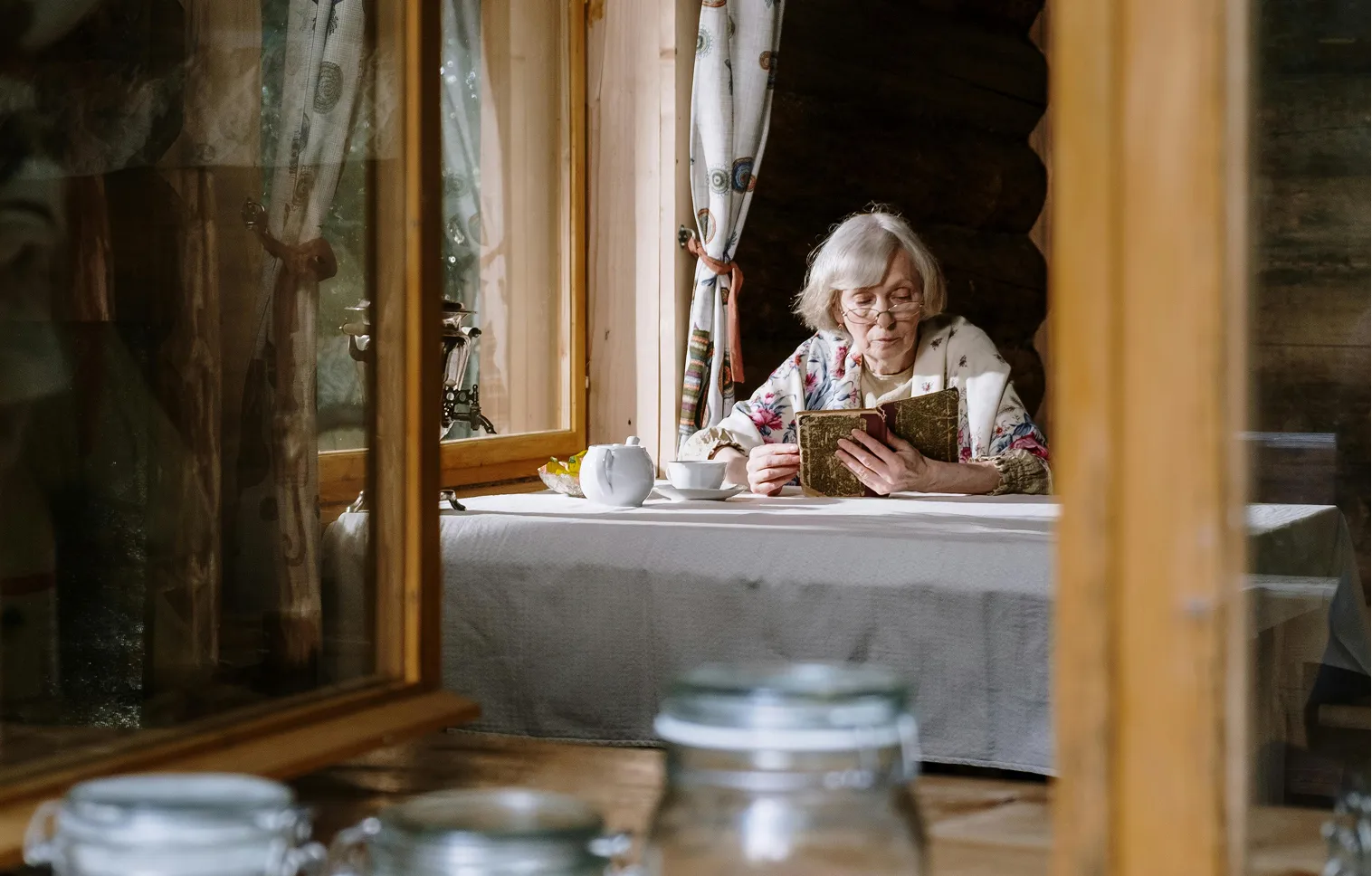 An elderly woman with gray hair and glasses reading a book at a table with a teapot and cup in a cozy wooden room.