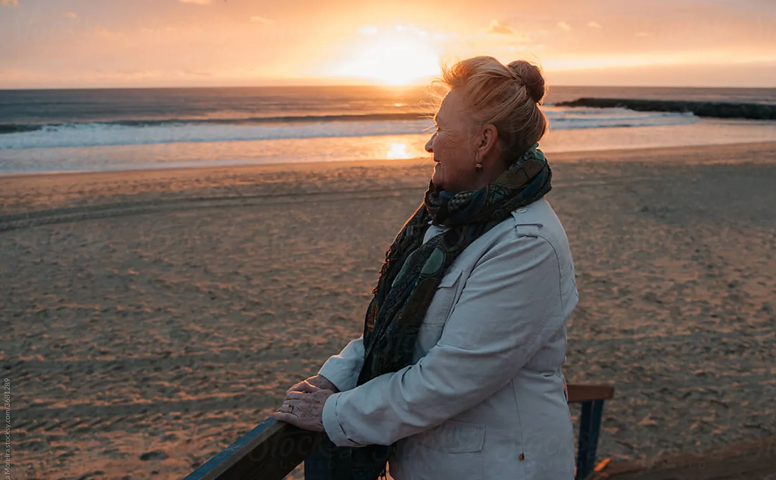 Elderly woman in a white coat and scarf gazing at the sunset over the ocean from a beach balcony.